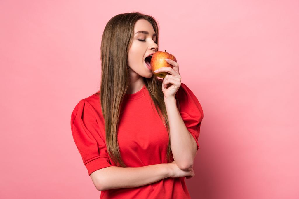 ελκυστική νεαρή γυναίκα τρώει μήλο με κλειστά μάτια σε ροζ - Φωτογραφία, εικόνα