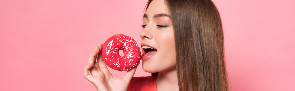 панорамний знімок привабливої дівчини, що їсть пончик із закритими очима ізольовано на рожевому
 - Фото, зображення
