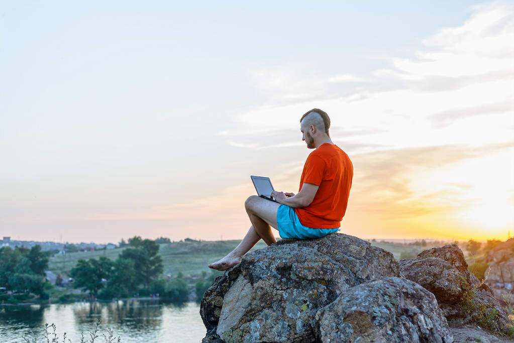 Freelancer working on a laptop while sitting on a cliff against the background of a sunset. - Photo, Image