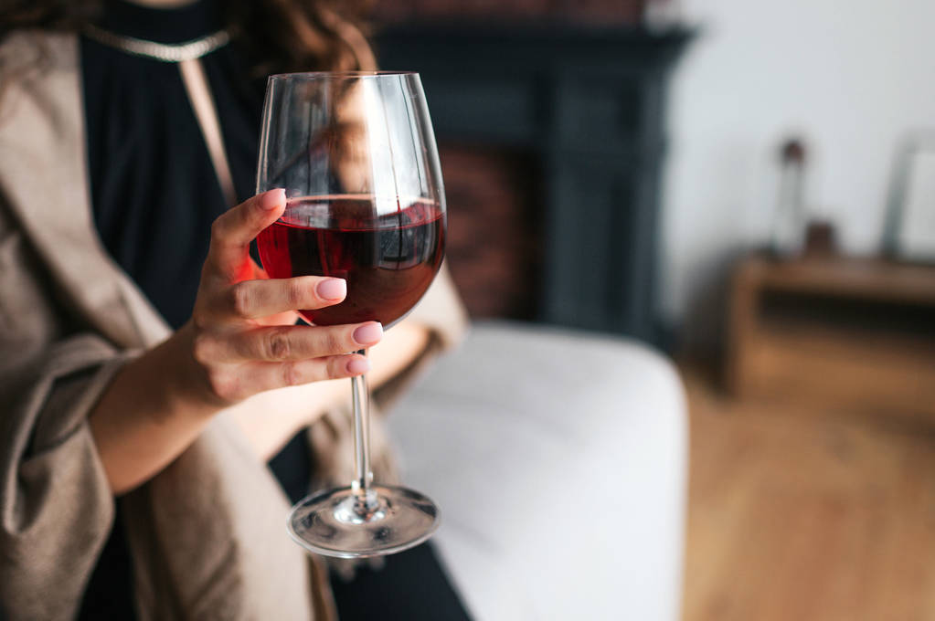 Pohled na ženské ruce držící sklenici červeného vína. Model nosí černé šaty a hnědou šálu. Žena v obýváku sama. - Fotografie, Obrázek