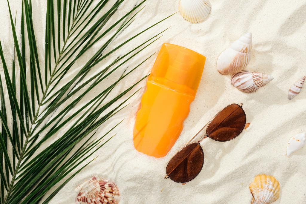 πορτοκαλί μπουκάλι αντηλιακού στην άμμο με κοχύλια, πράσινο φύλλο παλάμης και κομψά γυαλιά ηλίου - Φωτογραφία, εικόνα