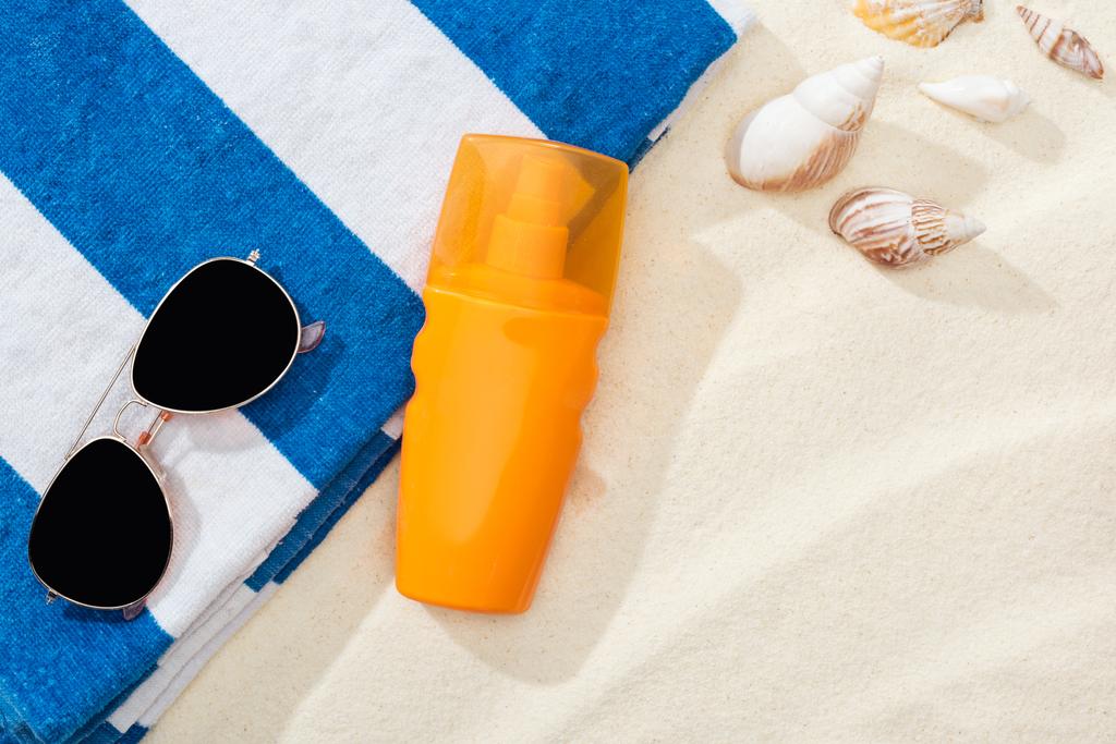 oranžová láhev opalovnice na písku s mušlemi, proužkovaný ručník a sluneční brýle - Fotografie, Obrázek