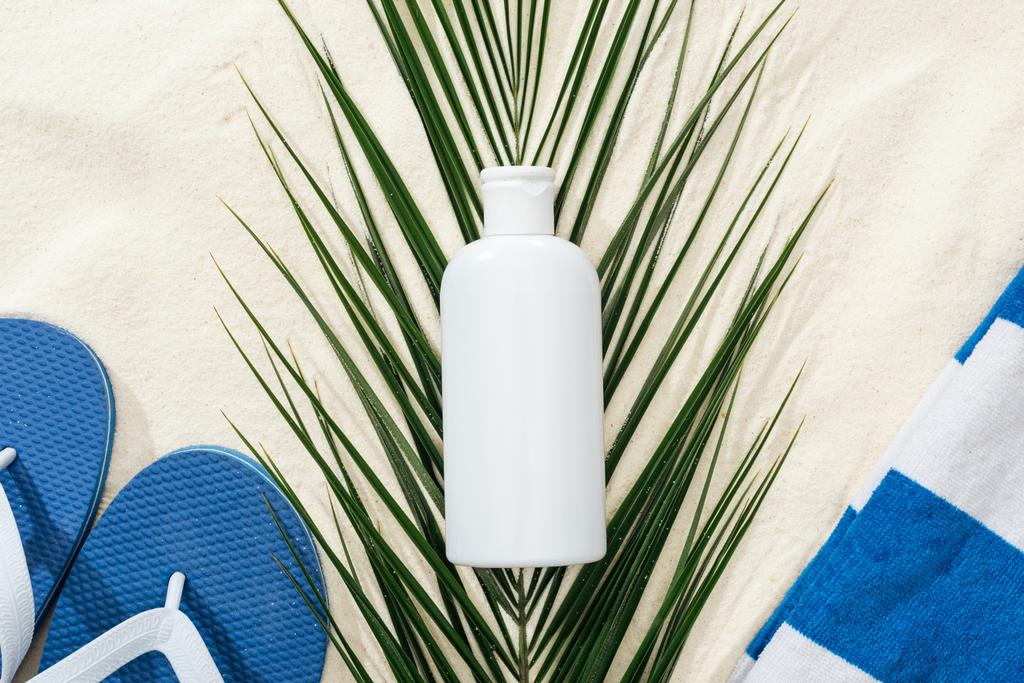 witte zonnebrandcrème lotion op groene Palm blad op zand met blauwe flip flops en handdoek - Foto, afbeelding