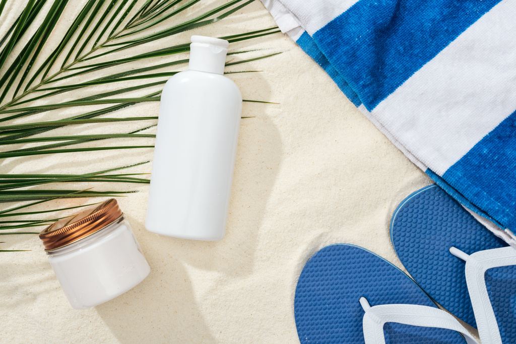balsam do opalania biały i krem w pobliżu zielonego liści palmowych na piasku z niebieskimi klapami i ręcznikiem - Zdjęcie, obraz