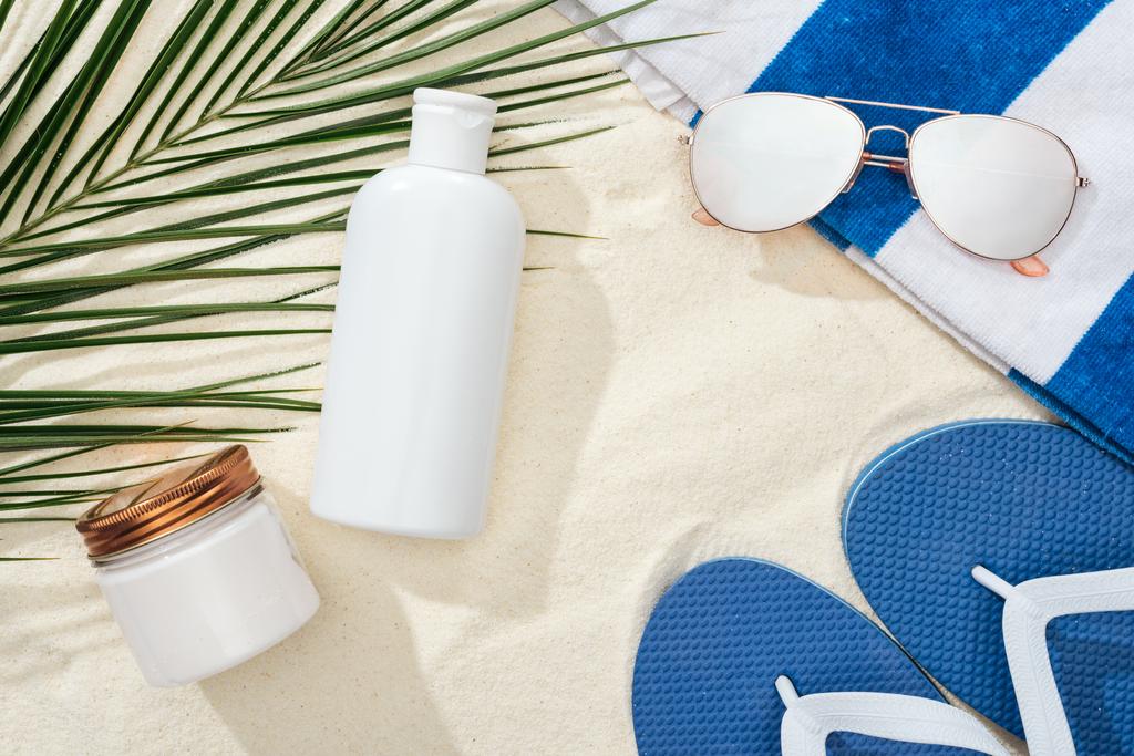 white sunscreen lotion and cream near green palm leaf on sand with blue flip flops, sunglasses and towel - Photo, Image