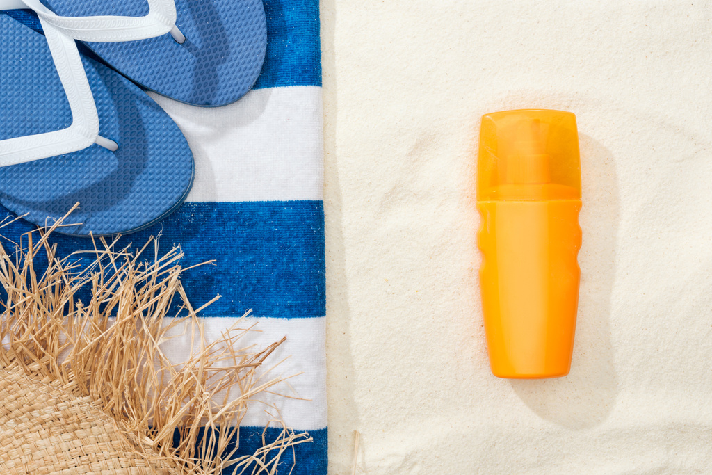 top view of sunscreen, straw hat, flip flops and striped blue and white towel on golden sand - Photo, Image