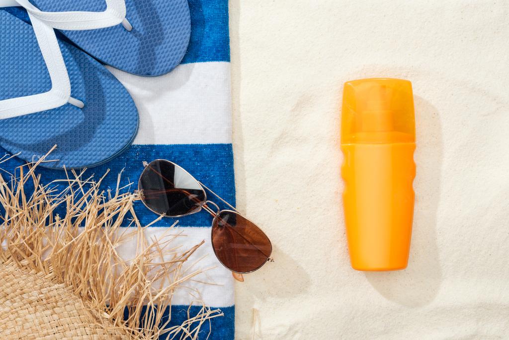 Top uitzicht van zonnebrandcrème, stro hoed, flip flops, zonnebril en gestreepte blauwe en witte handdoek op goudkleurig zand - Foto, afbeelding