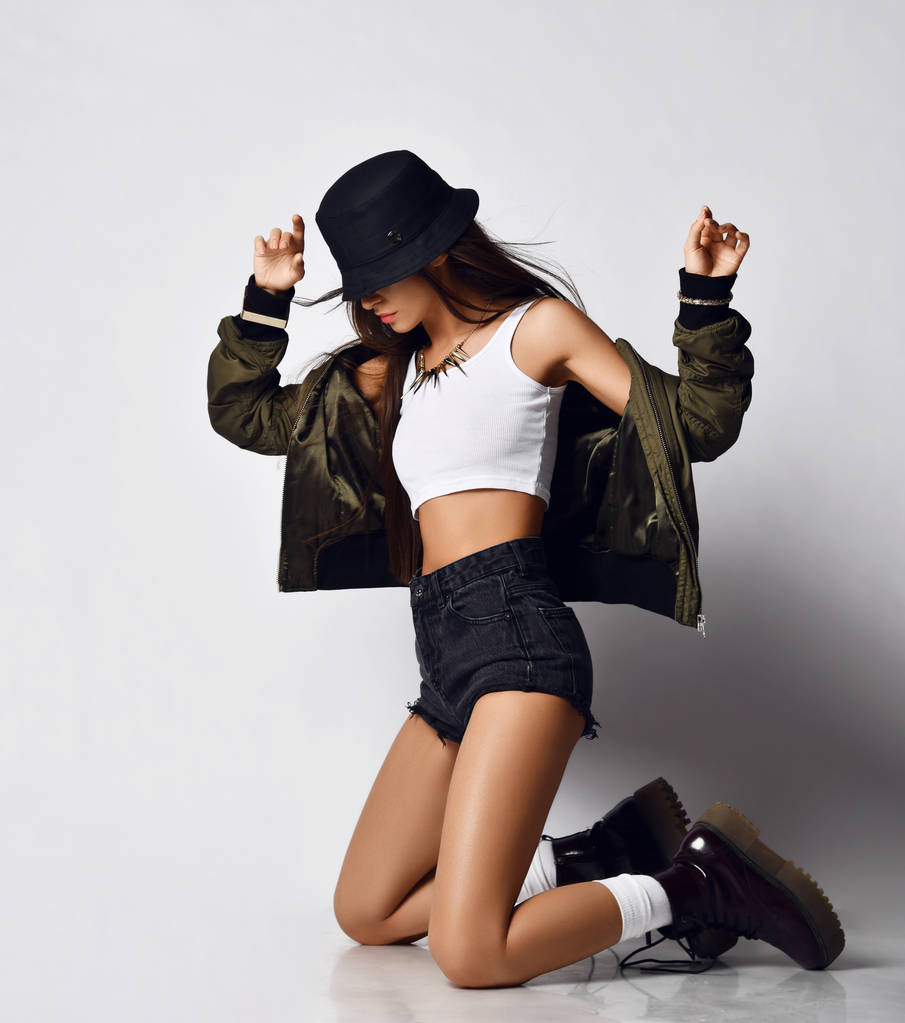 Young beautiful hipster girl sitting on her knees full body in shorts brown spring jacket modern stylish boots and hat  - Photo, Image