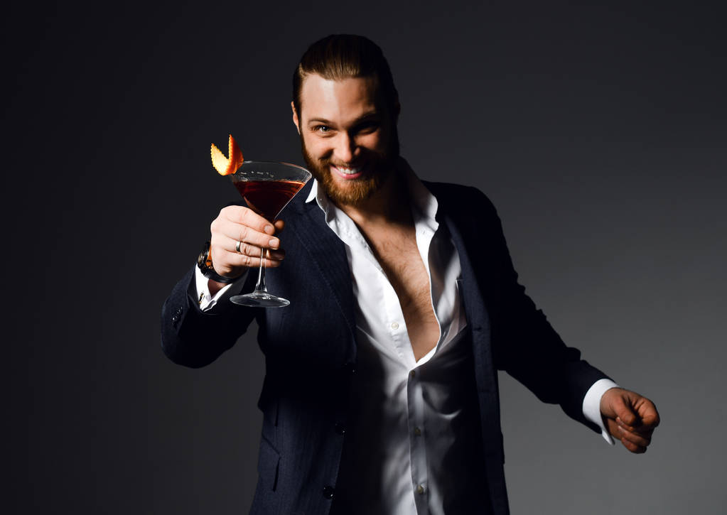 fröhlicher moderner bärtiger Mann im aufgeknöpften Hemd auf der Party bietet an, einen roten Cocktail mit Orange im Martini-Glas zu probieren - Foto, Bild