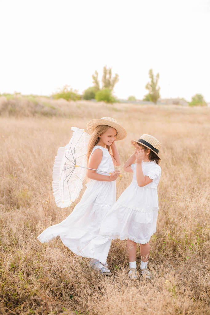 Carino sorelline con i capelli biondi in un campo estivo al tramonto in abiti bianchi con un cappello di paglia
 - Foto, immagini