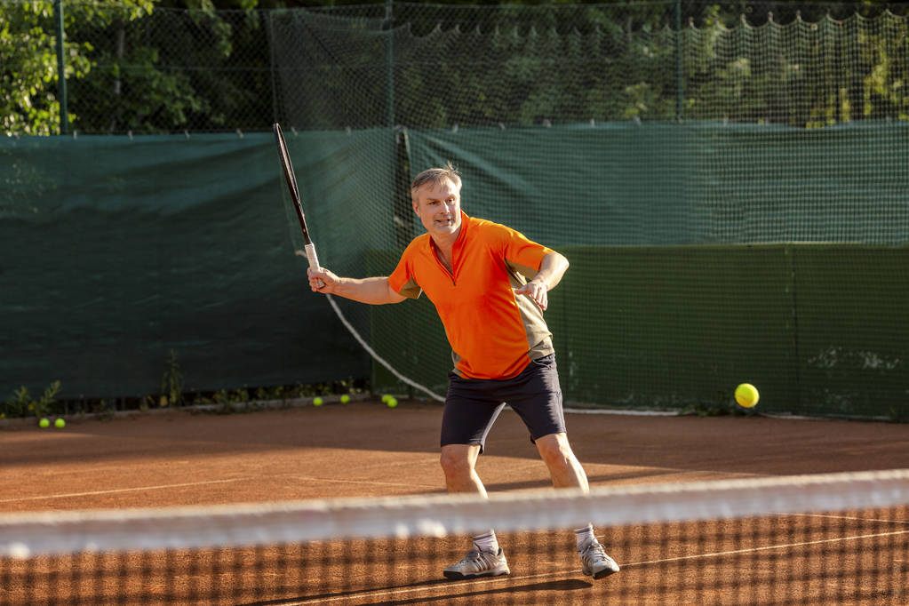 Mężczyzna w średnim wieku gra w tenisa na dworze z naturalną powierzchnią ziemi w słoneczny letni dzień. - Zdjęcie, obraz