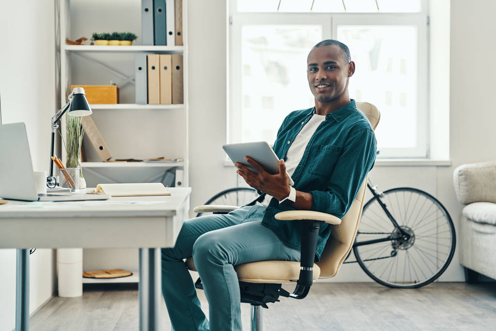 Ideoiden laittamista johonkin todelliseen. Komea nuori afrikkalainen mies paidassa työskentelee digitaalisella tabletilla istuessaan toimistossa
 - Valokuva, kuva