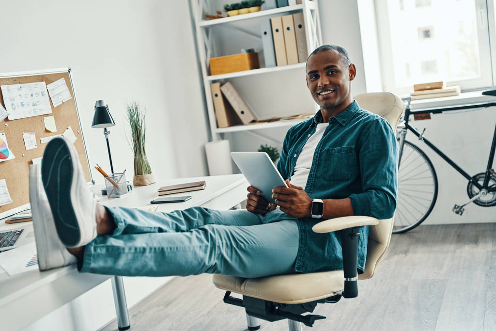 Sebevědomý manažer. Pohledný mladý Afričan v košili a při sezení v kanceláři se usmívá - Fotografie, Obrázek
