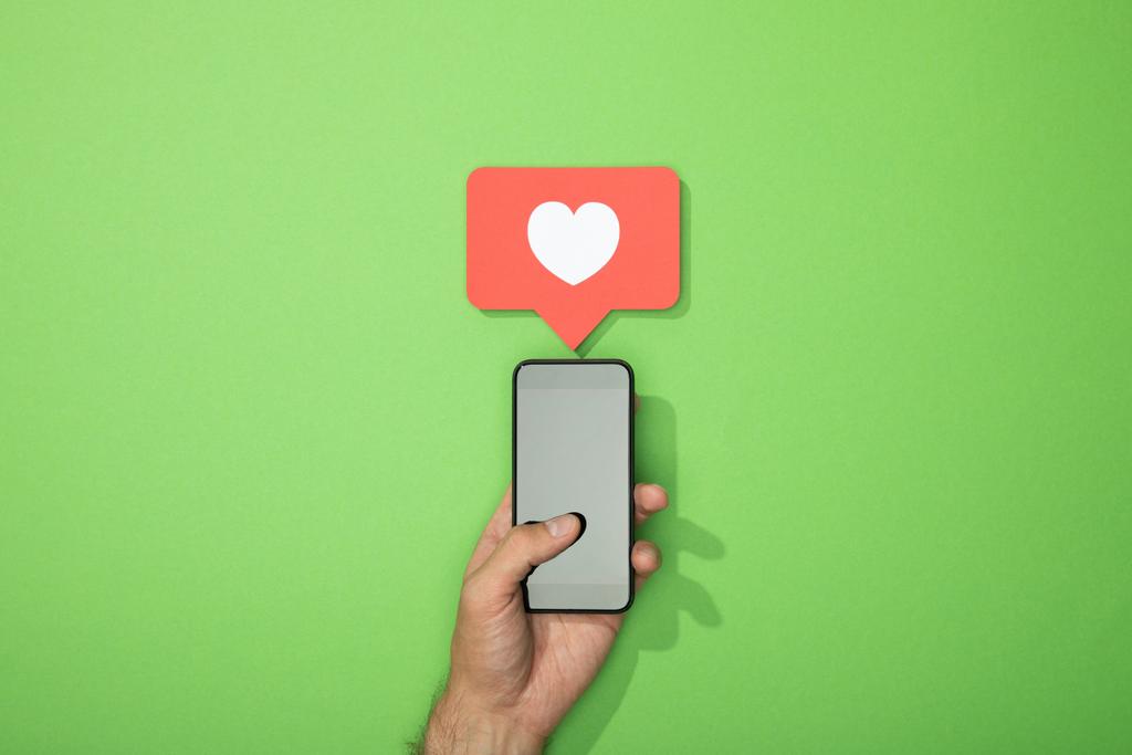 przycięte widok człowieka trzyma smartphone z białym sercem w ikonie powyżej gadżet na zielono - Zdjęcie, obraz