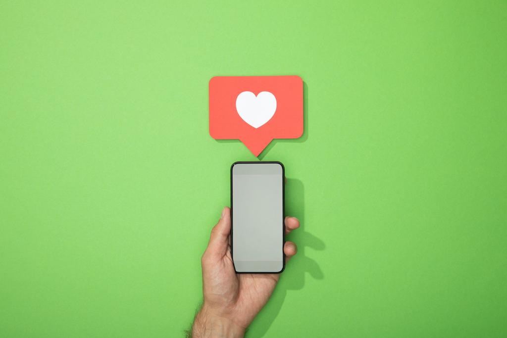 abgeschnittene Ansicht eines Mannes, der ein Smartphone mit Herz in rotem Symbol über einem Gadget auf Grün hält - Foto, Bild