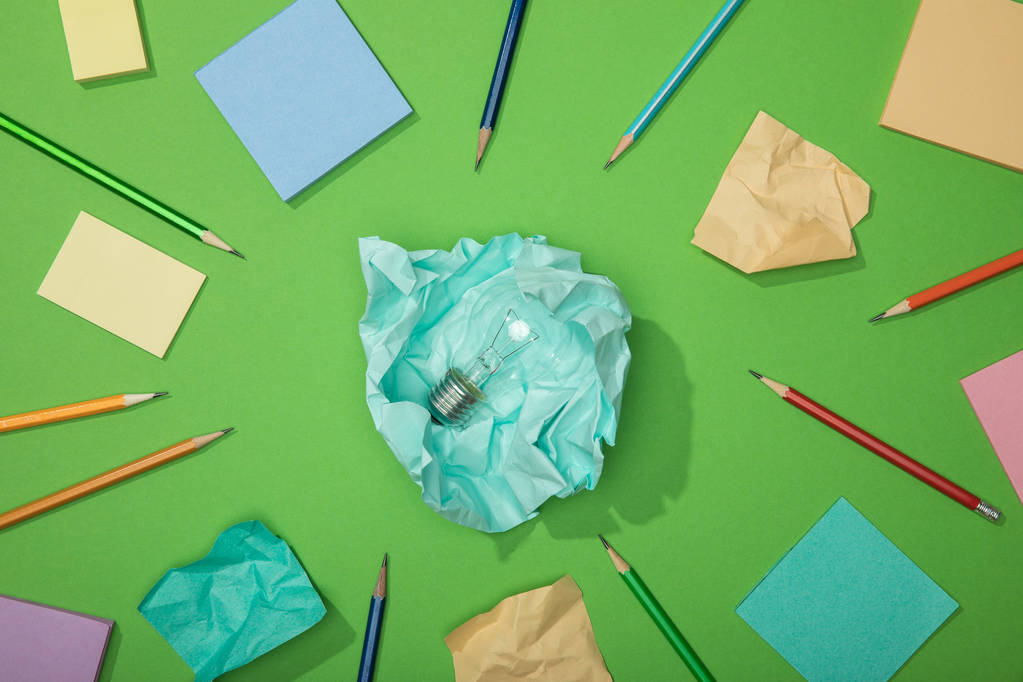 верхний вид лампочки на кусочке скомканной бумаги рядом с разбросанной бумагой и карандашами на зеленом
 - Фото, изображение