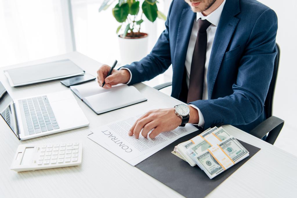 Ausgeschnittene Ansicht eines Mannes im Anzug, der in Notizbuch neben Geld und Laptop schreibt  - Foto, Bild