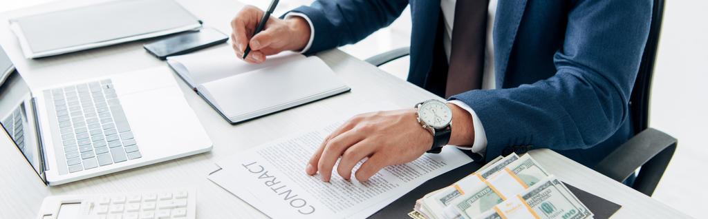 Panoramaaufnahme eines Mannes im Anzug, der in ein Notizbuch neben Laptop, Geld und Vertrag schreibt  - Foto, Bild