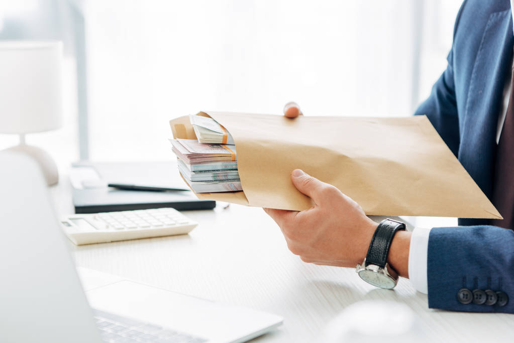 Ausgeschnittene Ansicht von Geschäftsmann mit Umschlag mit Geld und Laptop im Büro  - Foto, Bild