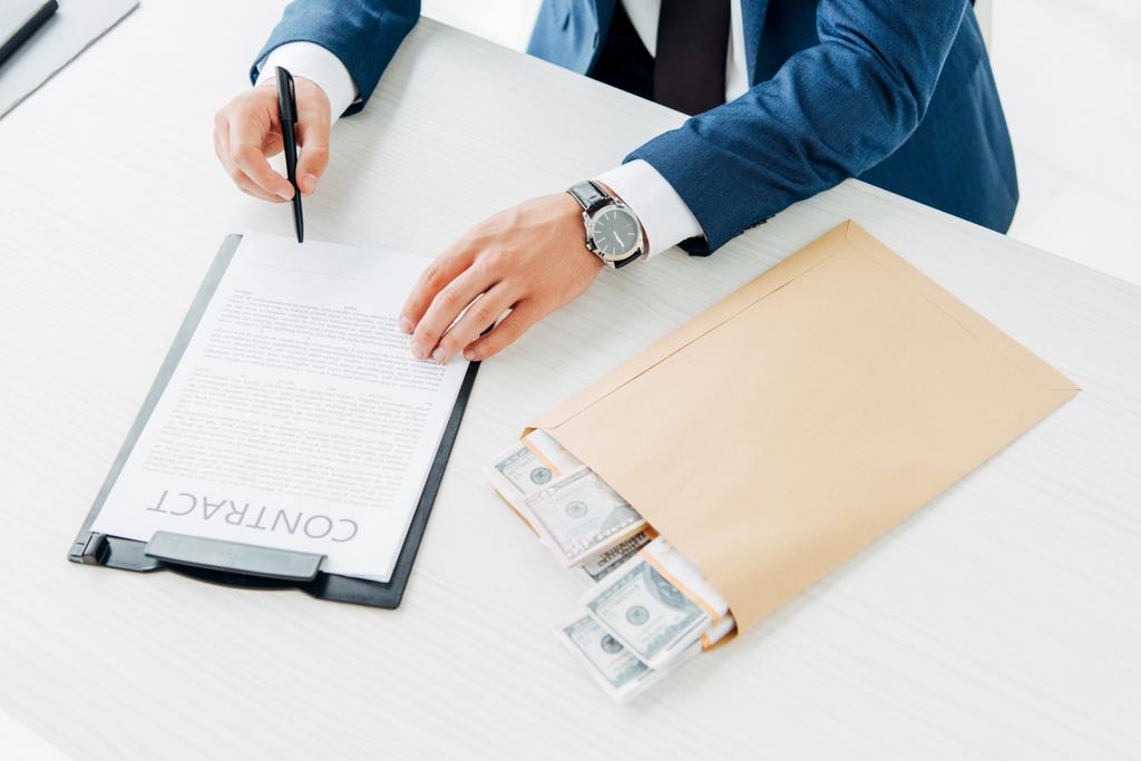 Κορυφαία όψη του επιχειρηματία που κρατάει στυλό ενώ υπογράφει σύμβαση κοντά σε φάκελο με χρήματα στο τραπέζι  - Φωτογραφία, εικόνα