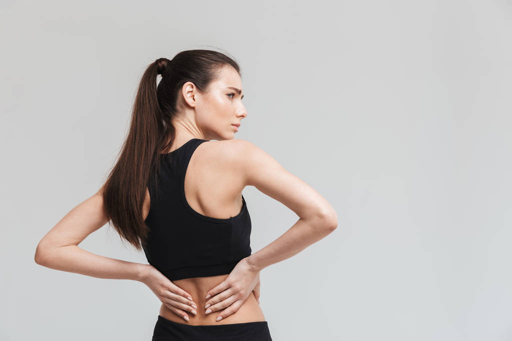 Bonita jovem triste esporte fitness mulher com dor nas costas isolado sobre fundo de parede cinza
. - Foto, Imagem