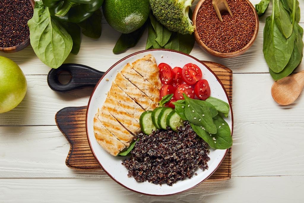 pohled na vařené quinoa s grilovaným kuřecím prsama a nakrájenou zeleninou na bílé desce na dřevěné sekací desce - Fotografie, Obrázek