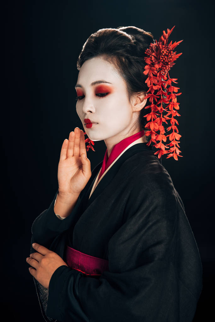 beautiful geisha in black and red kimono and flowers in hair with hand near face isolated on black - Photo, Image