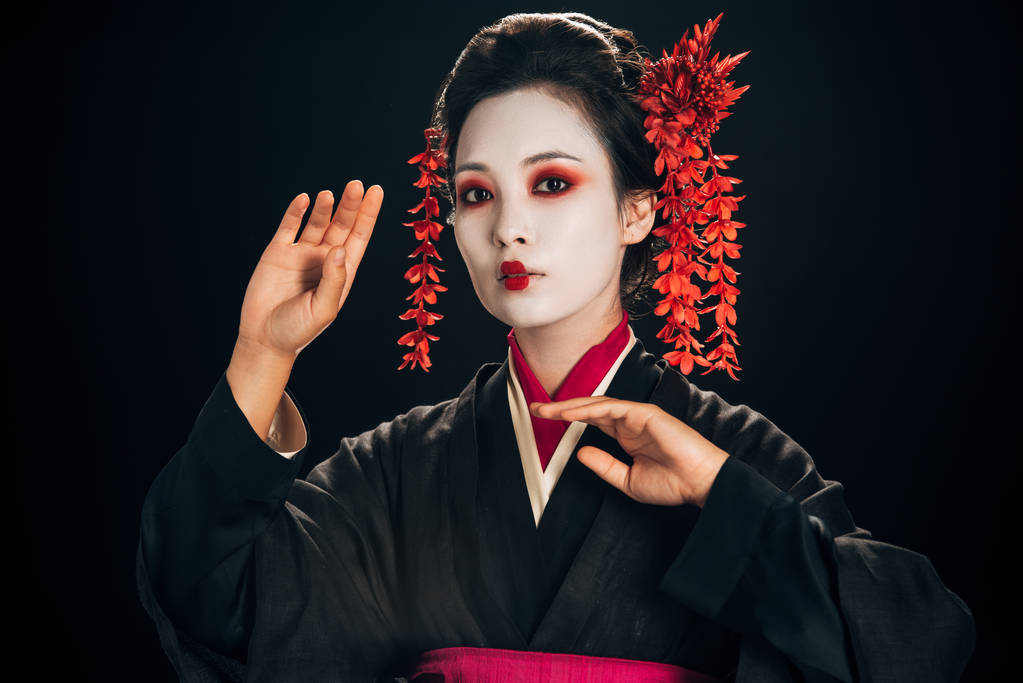 beautiful geisha in black and red kimono and flowers in hair gesturing isolated on black - Photo, Image