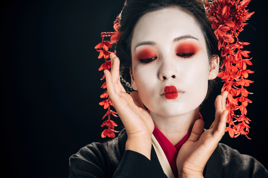 красивые гейши в черно-красном кимоно и цветы в волосах с руками возле лица, смотрящими вниз изолированы на черном
 - Фото, изображение
