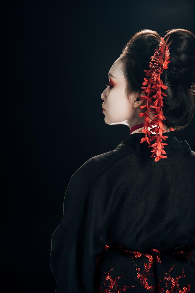 πίσω όψη της όμορφης γκέισα με μαύρο και κόκκινο κιμονό και λουλούδια στα μαλλιά κοιτάζοντας μακριά απομονωμένη σε μαύρο - Φωτογραφία, εικόνα