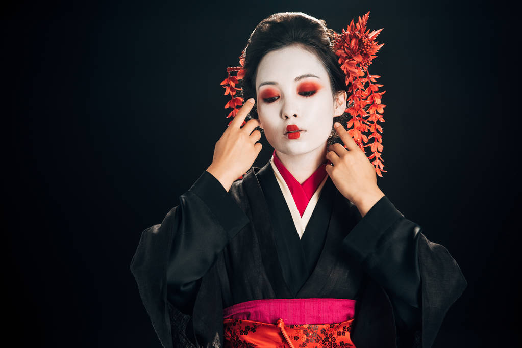 beautiful geisha in black and red kimono and flowers in hair touching face isolated on black - Photo, Image