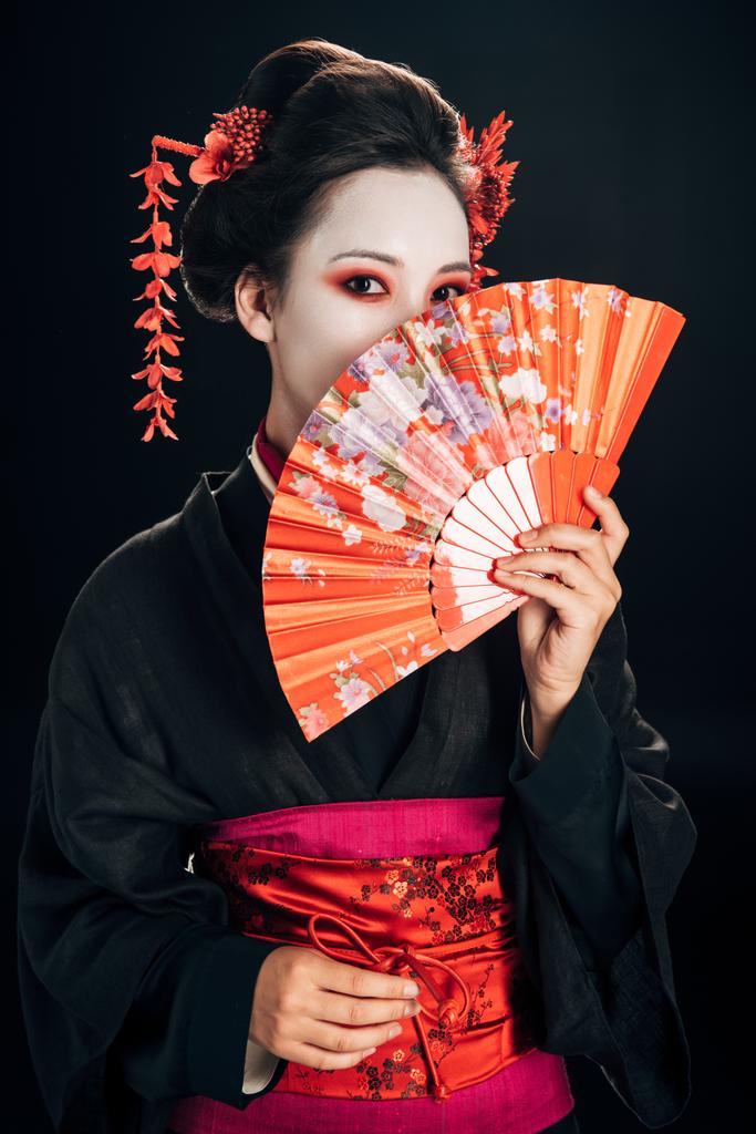geisha en kimono negro con flores rojas en el pelo ocultando la cara detrás de ventilador de mano tradicional aislado en negro
 - Foto, Imagen