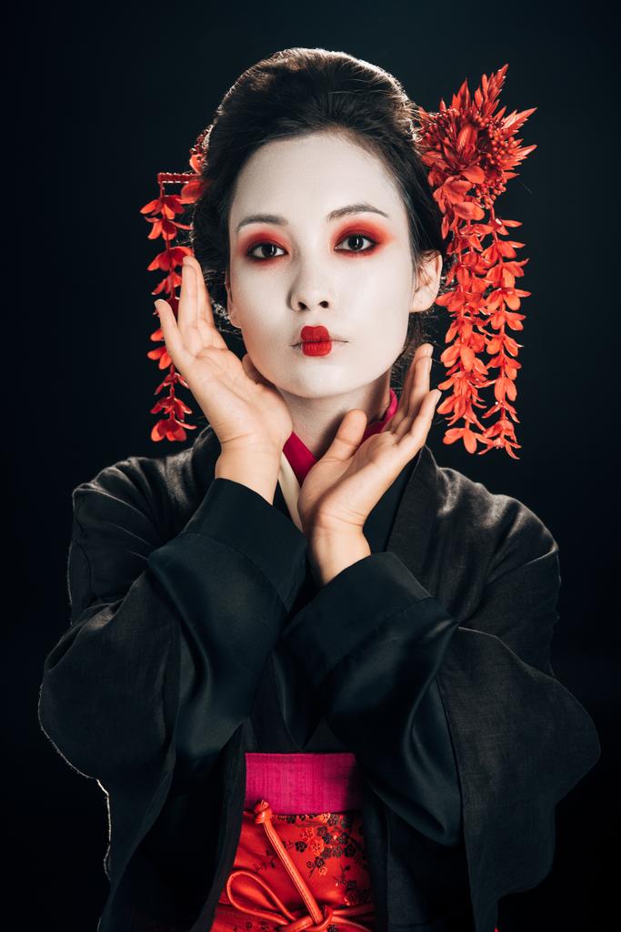 Geisha in zwarte en rode kimono en bloemen in haar met de hand in de buurt van gezicht geïsoleerd op zwart - Foto, afbeelding