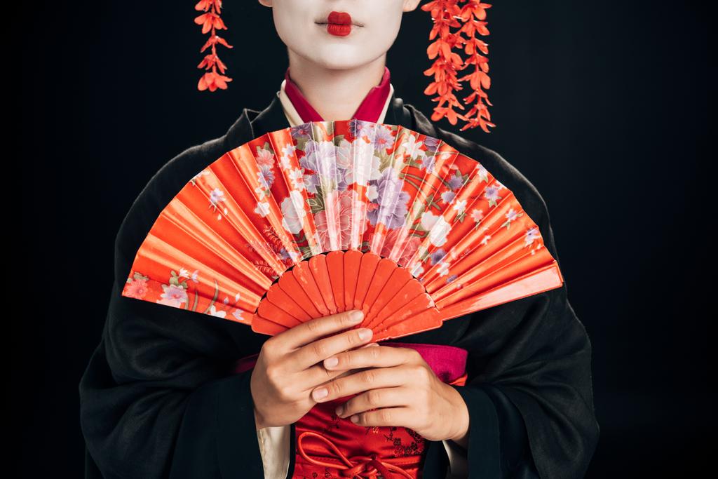 abgeschnittene Ansicht der schönen Geisha im schwarzen Kimono mit roten Blumen im Haar hält traditionellen Handfächer isoliert auf schwarz - Foto, Bild