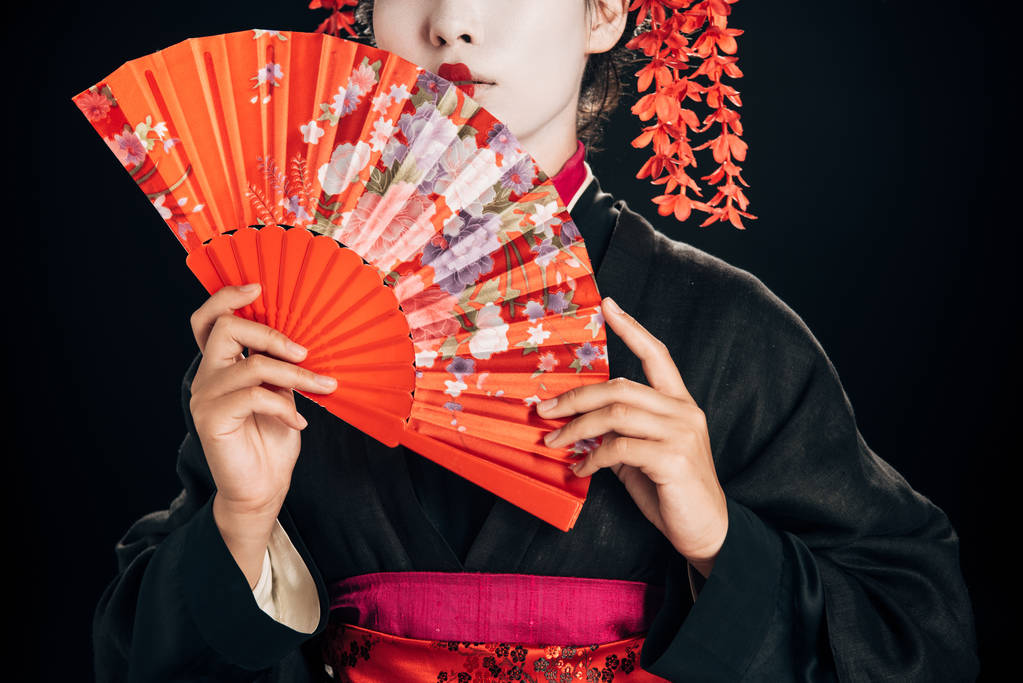 partial view of beautiful geisha in black kimono with red flowers in hair holding traditional hand fan isolated on black - Photo, Image