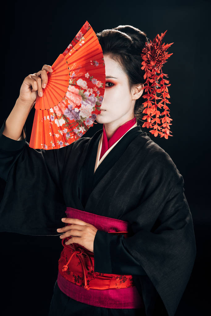 mooie Geisha in zwarte kimono met rode bloemen in haar schuil achter traditionele hand ventilator geïsoleerd op zwart - Foto, afbeelding