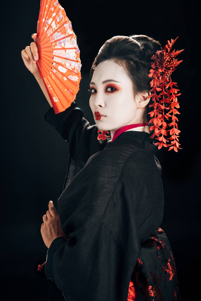 красивая гейша в черном кимоно с красными цветами в волосах жестикулируя традиционным ручным вентилятором, изолированным на черном
 - Фото, изображение
