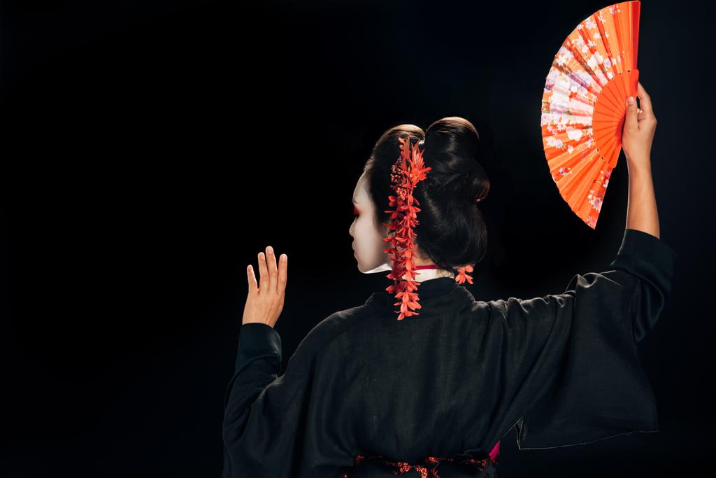 πίσω όψη της όμορφης γκέισα με μαύρο κιμονό με κόκκινα λουλούδια στα μαλλιά που κρατούν τον παραδοσιακό ανεμιστήρα χεριού και την προβολή που απομονώνεται σε μαύρο - Φωτογραφία, εικόνα