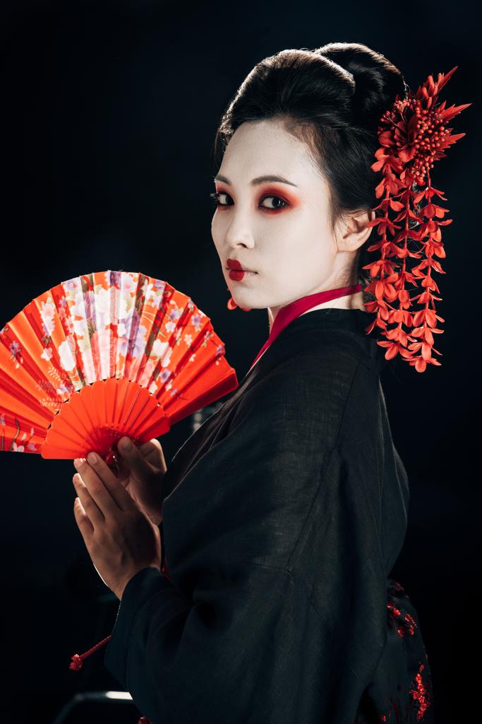 Gejša v černém kimonu s červenými květy ve vlasech držící tradiční ruční ventilátor izolovaný na černém - Fotografie, Obrázek