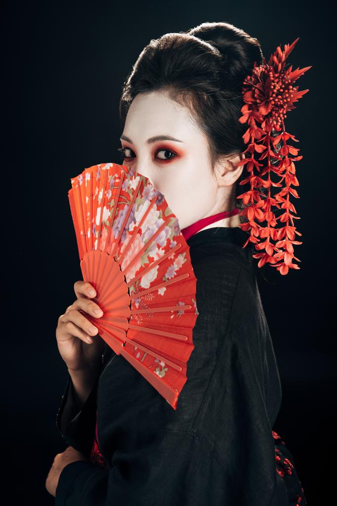 gyönyörű gésa fekete kimonó piros virágok a haj gazdaságban hagyományos kézi ventilátor izolált fekete - Fotó, kép