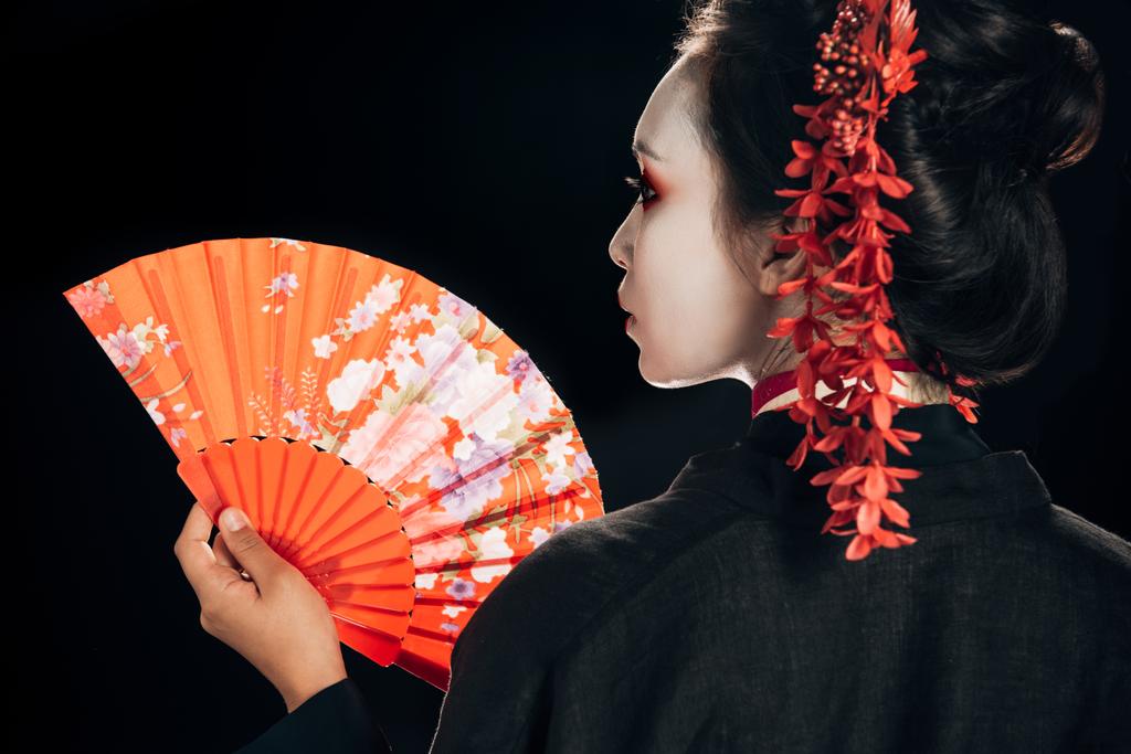 Rückseite der schönen Geisha im schwarzen Kimono mit roten Blumen im Haar, die den traditionellen Handfächer isoliert auf schwarz hält - Foto, Bild
