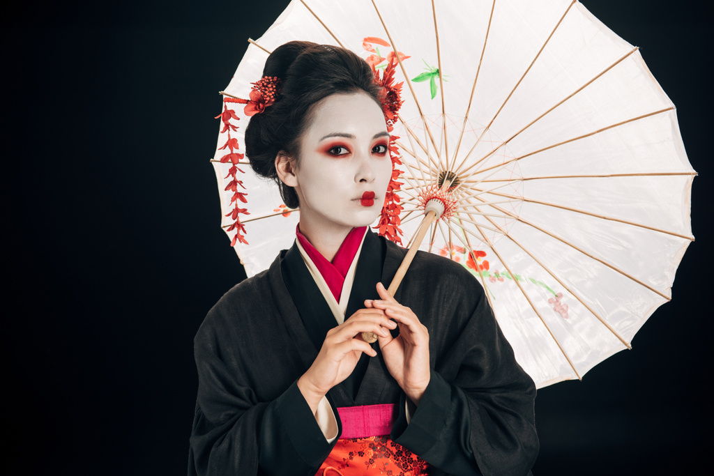 beautiful geisha in black kimono with red flowers in hair holding asian umbrella isolated on black - Photo, Image