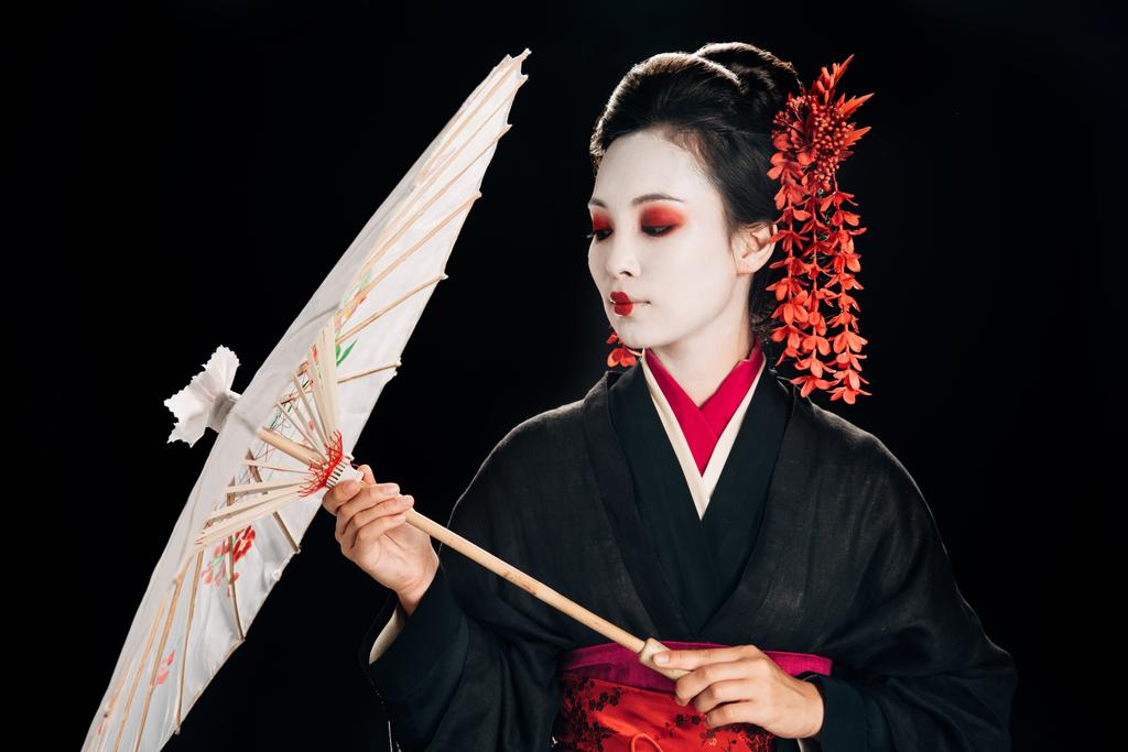 schöne Geisha im schwarzen Kimono mit roten Blumen im Haar, die auf den traditionellen asiatischen Regenschirm isoliert auf schwarz schauen - Foto, Bild