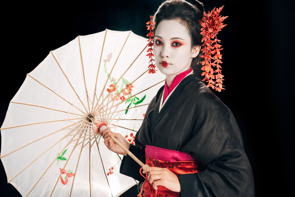 гейша в черном кимоно с красными цветами в волосах, держащих традиционный азиатский зонтик, изолированный на черном
 - Фото, изображение