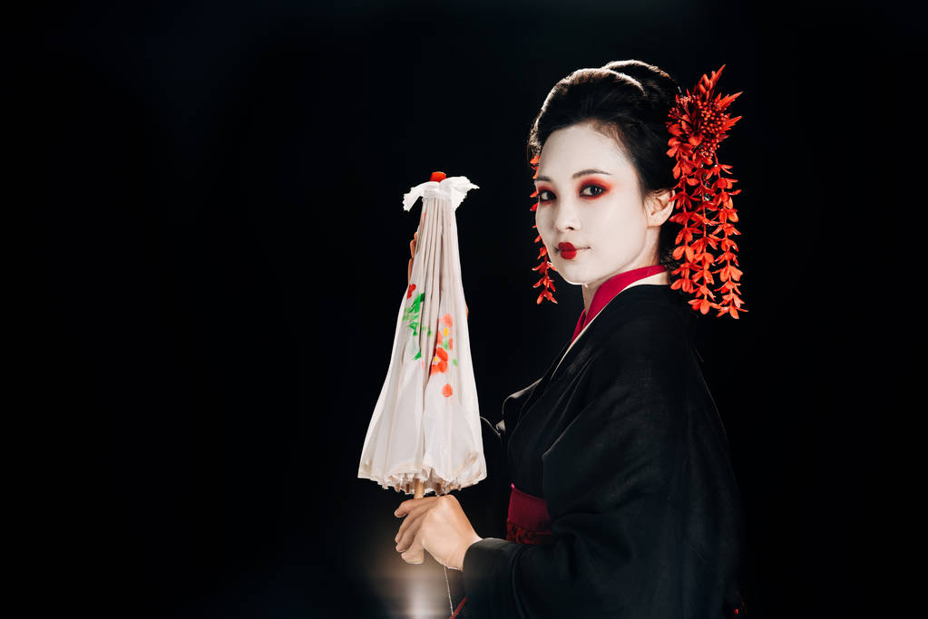 Side άποψη της γκέισα σε μαύρο κιμονό με κόκκινα λουλούδια στα μαλλιά κρατώντας παραδοσιακή ασιατική ομπρέλα απομονωθεί σε μαύρο - Φωτογραφία, εικόνα