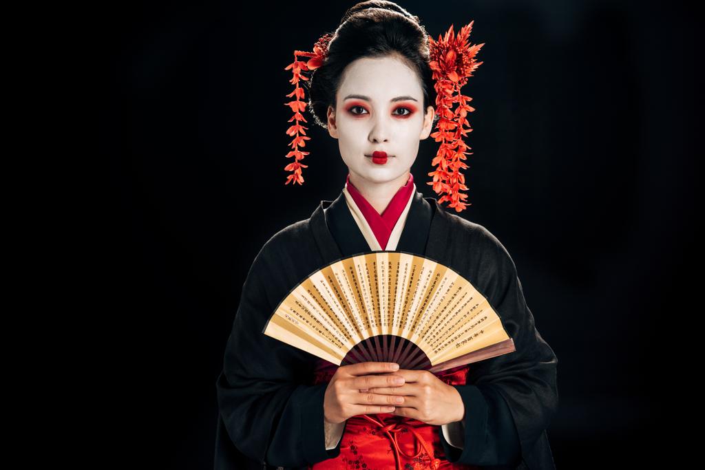 黒い着物の芸者は、黒で隔離された伝統的なアジアのハンドファンを持つ髪に赤い花を持つ - 写真・画像