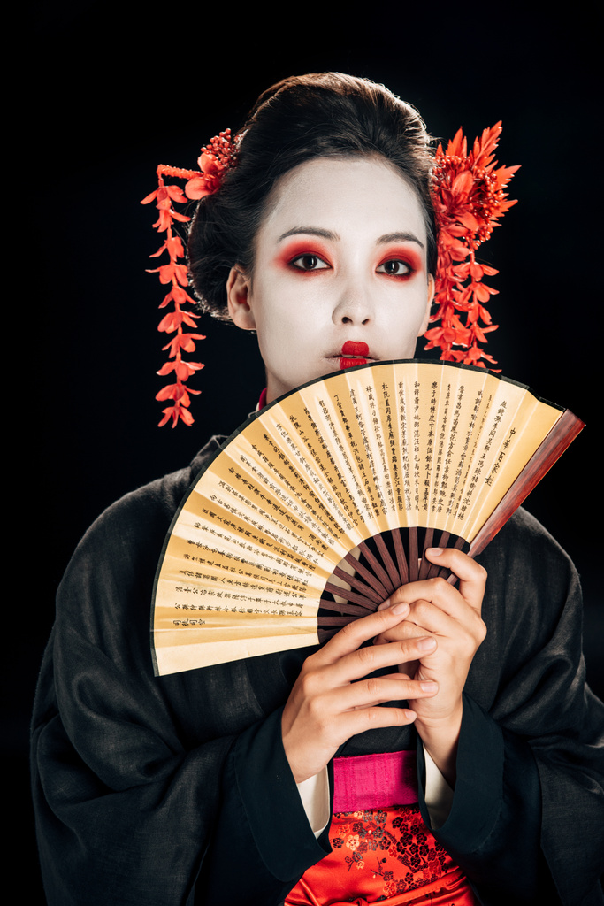 junge Geisha im schwarzen Kimono mit roten Blumen im Haar, die einen traditionellen asiatischen Handfächer isoliert auf schwarz hält - Foto, Bild