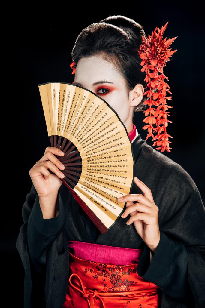 γκέισα με μαύρο κιμονό με κόκκινα λουλούδια στα μαλλιά και σκοτεινό πρόσωπο κρατώντας παραδοσιακό ασιατικό χέρι ανεμιστήρα απομονώνεται σε μαύρο - Φωτογραφία, εικόνα