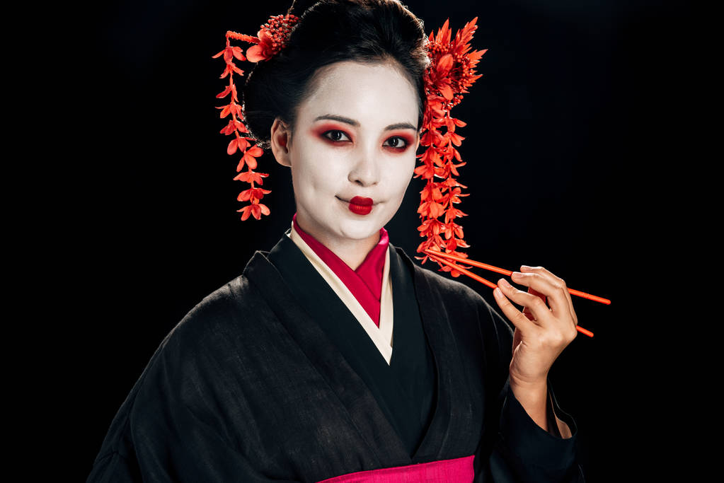 usmívající se krásná gejša v černém kimonu s červenými květy ve vlasech s hůlky, izolované na černém - Fotografie, Obrázek