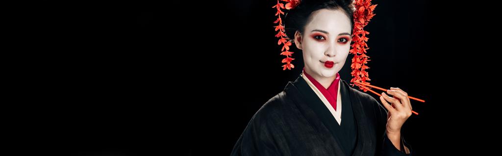 belle geisha souriante en kimono noir avec des fleurs rouges dans les cheveux tenant des baguettes isolées sur noir, plan panoramique
 - Photo, image